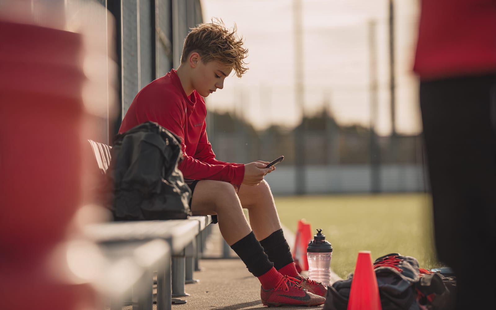 A young player on a bench checking his phone, kit bag and cleats beside him