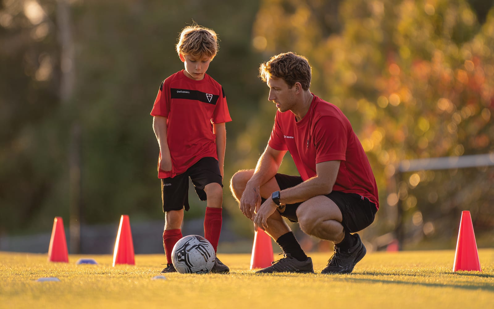 A coach kneeling beside a young player, working on his footwork at the ball